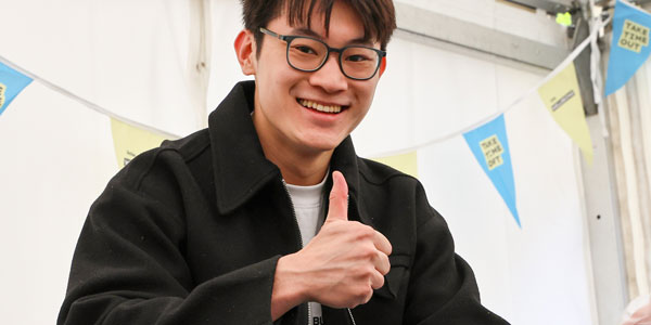 A student smiling and holding a thumbs up at the camera while they wait for a fresh smoothie to be made