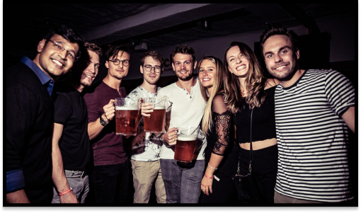 A photograph of a group of students in a bar, holding drinks and smiling at the camera